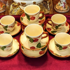 May include: A set of eleven cream-colored teacups and saucers. Each cup and saucer is decorated with a red cherry and green leaf design. The cups have brown handles and are arranged on a burgundy surface.