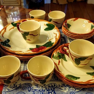 Puede incluir: Un juego de vajilla de cerámica color crema con un diseño de frutas. El juego incluye platos, cuencos y tazas con acentos de frutas y hojas rojas y verdes. Los bordes están adornados con un color marrón rojizo.