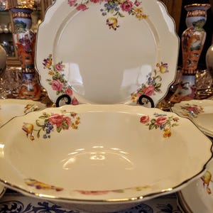 May include: Cream-colored porcelain serving dishes with a floral rose pattern. The set includes a large platter and a bowl, both with scalloped edges and gold trim. The floral design features pink, yellow, and blue flowers.