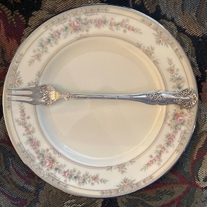 May include: A silver fork with an ornate design rests on a white plate with a floral pattern. The plate has a gold rim and is set on a patterned fabric.