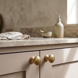 May include: Two round, brushed brass cabinet knobs are mounted on light beige, flat-panel kitchen cabinet doors. The knobs feature a smooth, circular face and a short, cylindrical base, providing a metallic contrast to the neutral cabinetry.