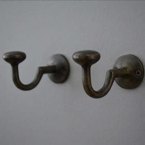 Könnte beinhalten: Zwei Wandhaken aus Metall mit einem dunklen, gealterten Messing-Finish. Jeder Haken verfügt über einen abgerundeten Knopf an der Oberseite, einen geschwungenen, J-förmigen Körper und eine runde Basisplatte, die mit einer sichtbaren Schraube befestigt ist.