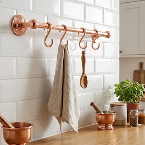 Puede incluir: Una barra de tubo de cobre pulido montada horizontalmente en una pared de azulejos blancos, con cinco ganchos a juego en forma de S. Un paño de cocina de lino beige y una cuchara de madera cuelgan de los ganchos para el almacenamiento.