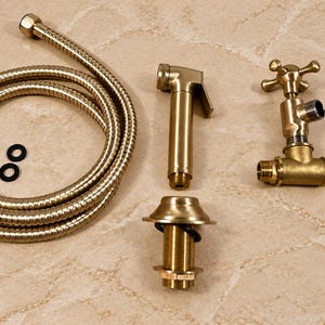 Könnte beinhalten: Ein Bidet-Sprühset in gebürstetem Gold, bestehend aus einem flexiblen Metallschlauch, einer Handbrause, einem Ventil mit Kreuzgriff, einer Montagebasis und zwei kleinen schwarzen Gummidichtungen mit metallischer Oberfläche.