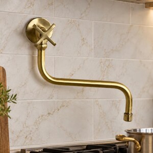 May include: A wall-mounted pot filler tap with a brushed brass finish. The fixture features a cross-handle design and a long, articulated spout that extends horizontally from the wall, positioned above a kitchen stove area.