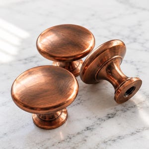 Puede incluir: Tres pomos redondos para armarios con acabado en cobre cepillado. Cada pomo presenta una superficie superior lisa y ligeramente abombada con una base cónica. El metal tiene un tono cálido marrón rojizo con finas vetas horizontales.