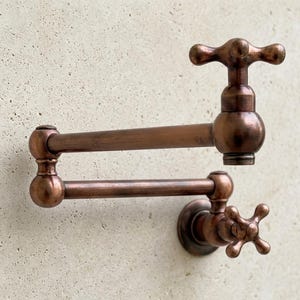 Könnte beinhalten: Ein wandmontierter Topffüller-Wasserhahn mit warmem, gebürstetem Kupfer-Finish. Diese bewegliche Armatur verfügt über zwei horizontale Arme, die durch ein zentrales Gelenk verbunden sind, ein Kreuzgriff-Ventil an der Basis und einen passenden Kreuzgriff.