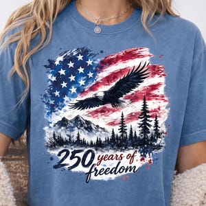 May include: Blue t-shirt featuring a patriotic design with an American flag, an eagle, mountains, and pine trees. The text "250 years of freedom" is printed below the graphic. The shirt is a comfortable, casual style.