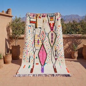 Puede incluir: Una alfombra rectangular de color crema con un diseño geométrico llamativo. La alfombra presenta una forma de diamante rosa central, rodeada de acentos coloridos y líneas negras. Tiene una franja en el borde inferior.