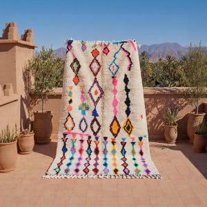 Puede incluir: Una alfombra rectangular de color crema con un vibrante patrón geométrico. El diseño presenta formas de diamantes y zig-zag coloridos en tonos de rosa, naranja, azul y amarillo. La alfombra tiene un borde con flecos y se exhibe al aire libre.