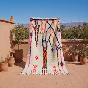 Könnte beinhalten: Ein weißer Teppich mit einem farbenfrohen geometrischen Design. Der Teppich weist vertikale Linien in Pink, Orange, Gelb und Blau mit schwarzen Akzenten auf. Der Teppich wird im Freien auf einer Terrakotta-Oberfläche mit Pflanzen und Bergen im Hintergrund präsentiert.