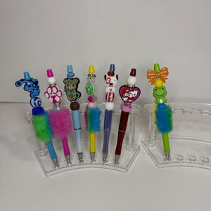 May include: A selection of decorative pens with diverse toppers, such as a paw print and a cartoon dog. The pens feature colourful fur accents and are displayed in a clear plastic holder. These pens are designed for writing.