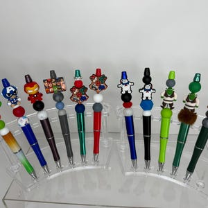 Peut inclure: Une collection de onze stylos présentés sur un support en acrylique transparent. Chaque stylo comporte un embout unique représentant un personnage ou un emblème, incluant une tortue, un super-héros et des écussons, avec des perles colorées.