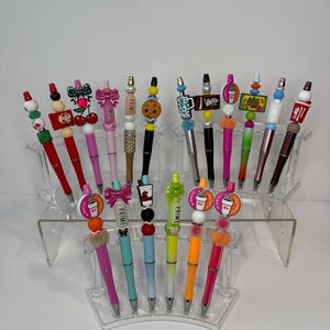 Könnte beinhalten: Eine Sammlung dekorativer Stifte mit verschiedenen bunten Aufsätzen, darunter Schleifen, Kekse und Kaffeetassen. Die Stifte werden auf transparenten Acrylständern ausgestellt. Die Stifte haben eine Vielzahl von Farben, darunter Rosa, Rot, Schwarz und Grün.