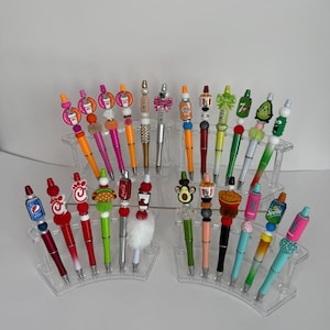 May include: A collection of twenty-one decorative pens arranged on three clear acrylic tiered stands. Each pen features a unique topper, including miniature food and beverage brand logos, colourful beads, fuzzy pom-poms, and various themed charms.