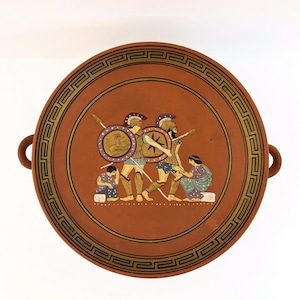 May include: A terracotta-colored ceramic plate with a detailed illustration of ancient Greek warriors. The plate features a decorative border and two small handles. The warriors are depicted with shields, spears, and helmets, in a scene of battle.