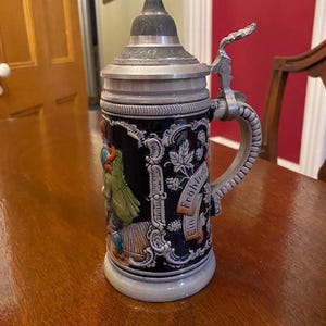 May include: A dark blue ceramic beer stein with a pewter lid and handle. The stein features a relief design of a person in green clothing and a banner with the text Ein Fröhliches. The lid has a pointed top and ornate metal detailing.