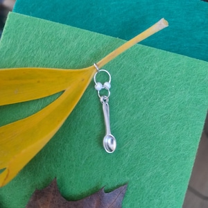 May include: A silver spoon charm earring with a small pearl accent hangs from a silver hoop. The earring is displayed on a green felt background with a yellow leaf and a brown leaf.