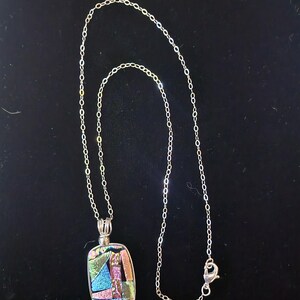 May include: A silver necklace with a rectangular pendant. The pendant features a colorful, abstract design with shades of blue, pink, green, and gold. The chain is delicate and has a lobster clasp.