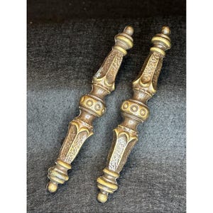 May include: A pair of antique bronze drawer pulls with intricate designs. Each pull has a textured handle, decorative circles, and rounded ends. These vintage pulls are likely for furniture restoration, with a classic aesthetic.