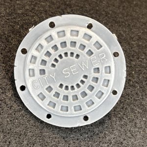 Puede incluir: Una tapa de alcantarilla en miniatura gris y circular con las palabras "CITY SEWER" en relieve en su superficie. La tapa presenta un patrón de rejilla y varios agujeros espaciados uniformemente alrededor del perímetro.