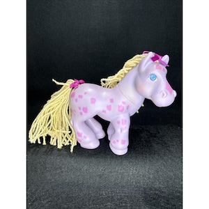 May include: A lavender toy pony with a yellow mane and tail. The pony has pink spots and a pink bow on its head and tail. The pony has blue eyes and is standing on a black surface.