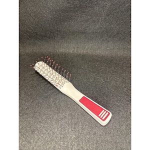 Pode incluir: Uma pequena escova de cabelo branca com um detalhe vermelho na pega. A cabeça da escova é retangular e preenchida com cerdas com pontas rosa. A pega tem uma secção vermelha com três linhas brancas.
