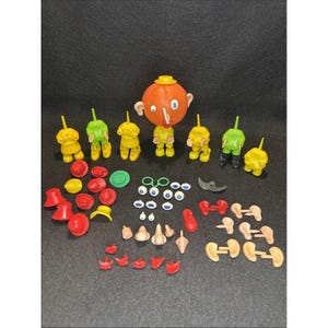 May include: A collection of vintage plastic toy parts, including orange heads, yellow and green figures, red hats, and various facial features like noses, eyes, and ears. The toys are arranged on a dark surface.