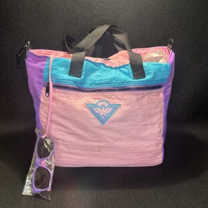 May include: A multi-colored tote bag with a pink, teal, and purple design. The bag has a black handle and a zipper pocket. A pair of purple sunglasses is attached to the bag.