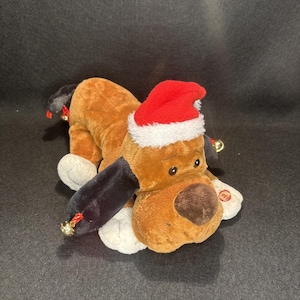 May include: A plush brown and tan dog wearing a red Santa hat with white trim. The dog has black ears, white paws, and a brown nose. Gold bells with red bows are attached to the ears. A red button is on the dog's paw.
