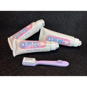 May include: Two white tubes of CPK toothpaste, each labeled 22 ml e (.75 fl. oz.) with pink and blue text and a tooth graphic, alongside a light purple plastic toothbrush with a white bristle head featuring a rectangular slot design.