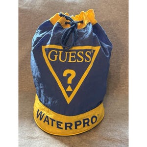 Puede incluir: Una bolsa azul y amarilla con cordón. Tiene la palabra "GUESS" en un triángulo amarillo y la palabra "WATERPRO" en amarillo. La bolsa tiene un cierre de cordón y está hecha de un material similar al nailon.