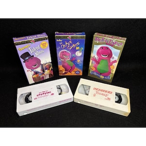 May include: A collection of Barney the Dinosaur VHS tapes. Titles include "Talent Show," "In Outer Space," and "Sing and Dance." The tapes feature colorful illustrations of Barney and related characters. The VHS tapes are white.