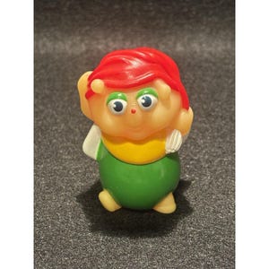 Könnte beinhalten: Eine Vintage-Plastikspielfigur eines lächelnden Charakters mit roten Haaren, grünen Augen und einem gelb-grünen Outfit. Das Spielzeug hat ein skurriles Design, mit erhobenen Armen und einem freundlichen Ausdruck.
