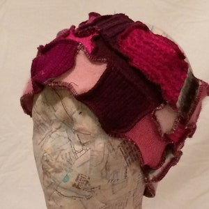 May include: A pink and purple knitted hat made from scraps of fabric. The hat is sewn together with a zig-zag stitch and has a unique, asymmetrical design.