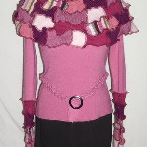 May include: A pink sweater with a ruffled neckline made from patchwork fabric in shades of pink, purple, and brown. The sweater has long sleeves with ruffled cuffs and a black skirt.