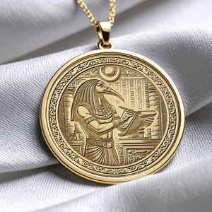 Puede incluir: Collar con colgante dorado con un relieve detallado de una deidad egipcia. El colgante circular representa una figura con cabeza de pájaro, escribiendo en un pergamino. El collar está sobre una cadena dorada, sobre un fondo de tela blanca.