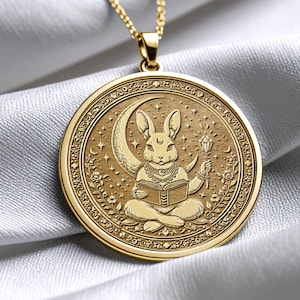 May include: A gold-coloured pendant necklace. The pendant features a detailed illustration of a rabbit seated in front of a crescent moon, holding a book and a staff. The round pendant has an ornate border and a delicate chain.