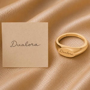 May include: A gold-toned signet ring with a smooth, polished surface featuring the engraved script text Dualora. Beside the ring is a square piece of light tan paper displaying the same brand name Dualora in a matching dark script font.