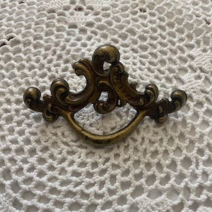 Puede incluir: Un tirador de cajón de latón vintage con un diseño decorativo y ornamentado. El tirador tiene una forma curva y detalles intrincados.