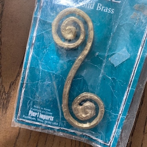 May include: Gold-colored metal drawer pull with a double spiral design. The pull is packaged in a plastic bag with a Pier 1 Imports label. The label reads "Pull Solid Brass Made in Korea Fabrique en Coree Pier 1 Imports Fort Worth, Texas 76161 USA."