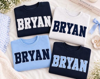 May include: Four crewneck sweatshirts featuring the name BRYAN in block lettering. One is navy with white text, one is light grey with navy text, one is light blue with navy text, and one is navy with light blue text.