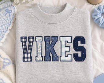 May include: A heather grey crewneck sweatshirt featuring the word VIKES in large, block-style letters. Each letter is filled with a different blue and white patterned fabric, including gingham, floral, and polka dot prints, with a dark blue border.