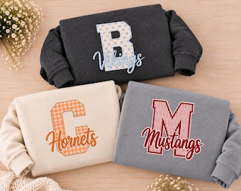 May include: Three crewneck sweatshirts in charcoal, cream, and heather grey. The charcoal sweatshirt features a blue floral letter B and the text Vikings. The cream sweatshirt has an orange gingham letter C and the text Hornets. The grey sweatshirt displays a red striped letter M and the text Mustangs.