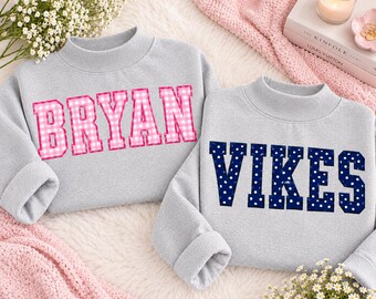 May include: Two light grey mock neck sweatshirts. The left sweatshirt features the name BRYAN in pink and white gingham block letters. The right sweatshirt features the word VIKES in navy blue block letters with white polka dots.