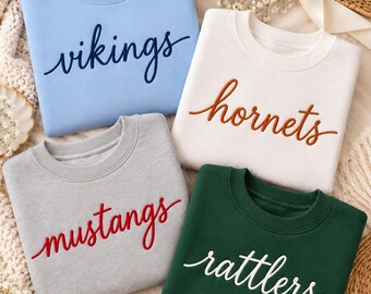 May include: Four crewneck sweatshirts in light blue, white, grey, and dark green. Each features cursive embroidery across the chest: dark blue text reading vikings, brown text reading hornets, red text reading mustangs, and white text reading rattlers.