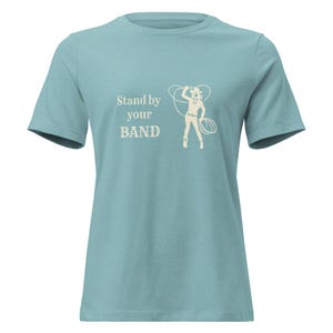 Pode incluir: Camiseta azul clara com o texto "Stand by your BAND" e um desenho creme de uma cowgirl com um laço. A camiseta tem mangas curtas e gola redonda.