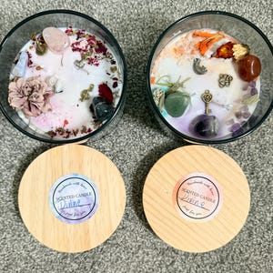 Può includere: Due candele rotonde in vetro con cera bianca, decorate con cristalli, fiori secchi e ciondoli in metallo. Ogni candela ha un coperchio in legno chiaro con un adesivo circolare che riporta la scritta "Handmade with love, SCENTED CANDLE, Divine, Just for you."