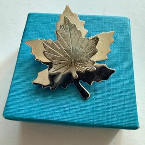 May include: A silver maple leaf brooch with intricate detailing, set on a textured turquoise box. The brooch has a three-dimensional design and a slightly aged appearance, possibly vintage. The box is a vibrant turquoise colour.