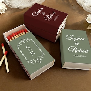 May include: Three decorative matchbox sets. One is open, revealing red-tipped matches. The boxes are in shades of green and burgundy, with white text that reads "Sophia & Robert" and the date "09.18.2026". The open box has the initials "S R".